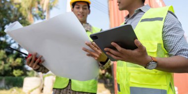 Endüstriyel bir ortamda lojistik ve konteyner yönetimine odaklanan, planlar ve tablet inceleyen güvenlik yelekleri ve miğferler üzerinde çalışan iki mühendisin kapatılması.