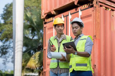 Güvenlik yeleği ve miğfer giyen iki mühendis, bir tablet ve planların lojistik planlarını incelerken, sanayi yönetimindeki işbirliğini vurguladılar..