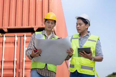 Güvenlik donanımlı iki mühendis, lojistik ve sanayi yönetimi üzerine yoğunlaşan bir nakliye konteynırının yakınındaki planları ve bir tableti inceliyor..
