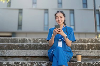 Moladan zevk alan bir hemşire kahve ve akıllı telefonuyla merdivenlerde oturup çağdaş bir tıbbi ortamda mutluluk saçar..