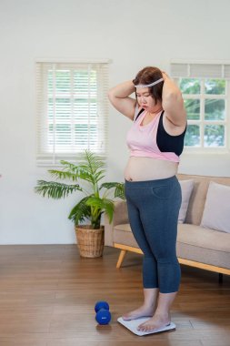 Büyük beden bir kadın kararlı bir tartıda duruyor, yanında halterlerle egzersiz yapmaya hazır, parlak bir yaşam alanındaki kilo verme yolculuğunu yansıtıyor..