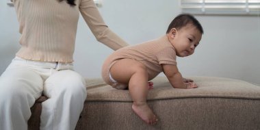 A baby attempting to crawl from a cozy sofa while a supportive parent gently assists them, creating a safe and loving atmosphere in their modern home. clipart