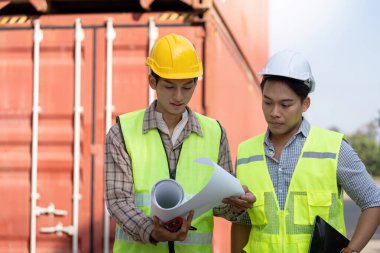 Güvenlik yeleği ve miğfer mühendisi, lojistik ve sanayi idaresini vurgulayan bir nakliye konteynırı yakınındaki planları ve planları gözden geçiriyor..