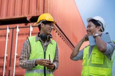 Güvenlik yelekleri ve kasklarındaki iki mühendis lojistik, el cihazları ve nakliye konteynırlarının yanında durup endüstriyel yönetimdeki takım çalışmalarını tartışırken bir ışık anını paylaşıyorlar..
