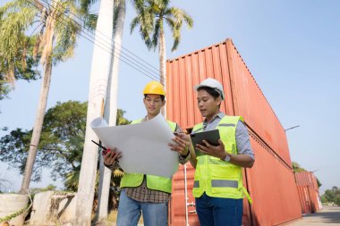 Güvenlik yelekleri ve miğferlerdeki iki mühendis, sanayi lojistik yönetimindeki işbirliğini vurgulayan nakliye konteynırlarıyla çevrili planları ve bir tableti dikkatle gözden geçiriyorlar..