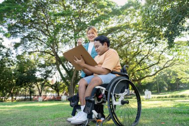 Tekerlekli sandalyedeki engelli genç bir sanatçı sanatını icra ediyor. Yeşillik dolu, güneşli bir parktaki destekleyici bir eğitmenden esinlenerek..