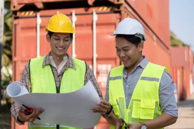 Güvenlik yeleklerinde çalışan iki profesyonel, planları incelerken, takım çalışması ve lojistik ve mühendislik alanlarında planlama yaparken gülümsüyor.