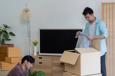 Two men engaged in unpacking boxes in their modern living room, with one sorting through items and the other smiling, surrounded by plants and a cozy ambiance. clipart