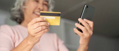 İnternetten alışveriş yapan yaşlı bir kadın, bir elinde kredi kartı, diğer elinde akıllı bir telefon, rahat bir evde keyif verici şeyler söylüyor..