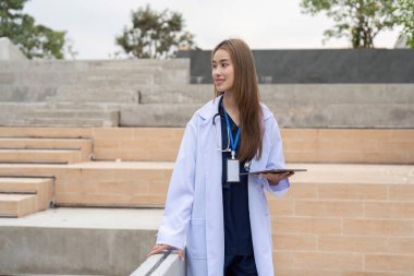 Düşünceli bir Asyalı kadın doktor elinde bir tablet ve steteskopla dışarıda duruyor. Çağdaş bir sağlık ortamında hasta danışmanlığına hazır..