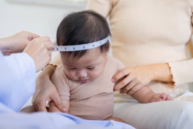 A pediatrician is measuring a babys head circumference using a tape measure, while the mother gently holds the infant in a medical clinic, monitoring growth. clipart