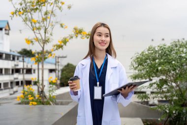 Kendine güvenen bir doktor elinde kahve fincanı ve tabletle dışarıda duruyor, hasta etkileşimlerine hazır, profesyonelliğin ve sağlık hizmetlerinin bir karışımını gösteriyor..