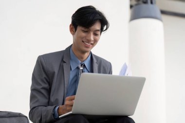 A young Asian businessman in a suit, smiling as he works on his laptop, showcasing a positive and productive atmosphere in a contemporary outdoor setting. clipart