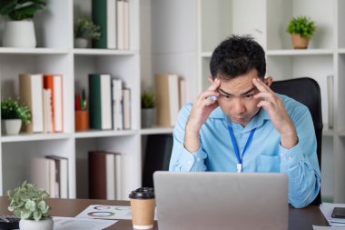 Asyalı bir işadamı yoğun bir şekilde bilgisayarına odaklanmış, çağdaş bir ofis ortamında masasında zorluklarla boğuşuyor..