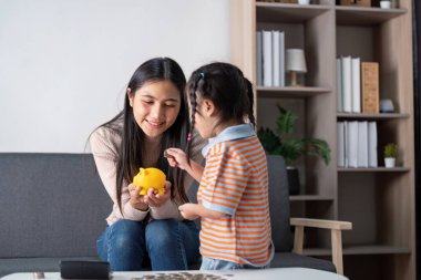 Çeşitli bir anne ve kızı, oturma odalarındaki sarı kumbarayı kullanarak birlikte para biriktirmeyi keşfederken bir neşe anlarını paylaşıyorlar..