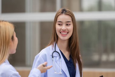 Beyaz önlüklü iki farklı kadın doktor bir sağlık merkezinin dışında hasta bakımı hakkında tartışıyorlar, sağlık hizmetlerinde işbirliğini vurguluyorlar..