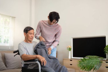 Düşünceli bir oğul, yaşlı babasının tekerlekli sandalyesine sıcak bir battaniye örterek rahat bir ev ortamında sevgi ve ilgiyi gösterir..