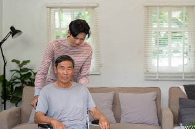 Şefkatli bir oğul, yaşlı babasına tekerlekli sandalyede yardım ediyor. Rahat bir evde sevgi ve aile desteği gösteriyor..