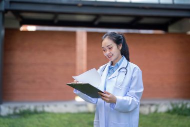 Beyaz önlüklü odaklanmış bir doktor, hasta belgelerini panoda incelerken gülümsüyor ve sağlık hizmetlerindeki kalite ve profesyonelliğe olan bağlılığını vurguluyor..