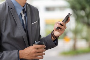 Takım elbiseli genç bir Asyalı işadamı elinde kahve fincanı tutarken akıllı telefonunu kullanıyor, işini ve günlük hayatını canlı bir açık hava ortamında harmanlıyor..