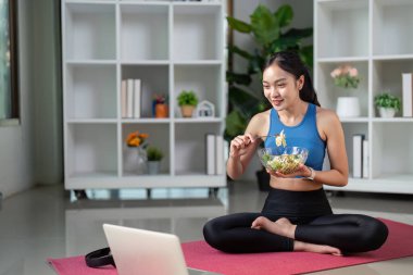 Spor kıyafetli bir kadın yoga minderinde besleyici bir salata yemekten hoşlanır. Dizüstü bilgisayarıyla yeni ve davetkar bir ortamda meşgul olur..