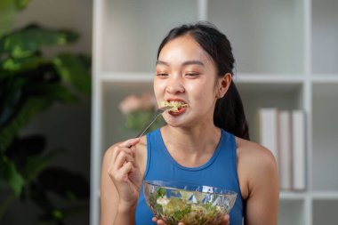 Çatal dolusu taze salatadan zevk alan bir kadın, kapalı bir ortamda mutlu bir şekilde gülümseyen, sağlıklı beslenme alışkanlıklarını teşvik eden bir kadın..