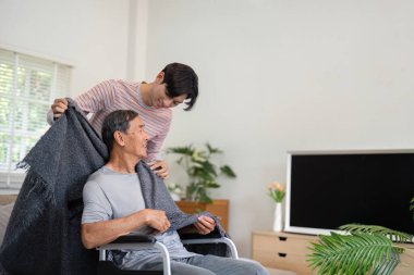 A son gently drapes a blanket over his elderly father sitting in a wheelchair, showcasing warmth and care in a comfortable home setting. clipart