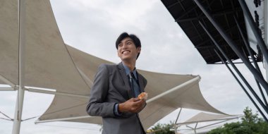 Takım elbise giymiş gülümseyen bir işadamı, açık havada büyük bir kumaşın altında sandviç tutuyor, mola sırasında neşeli bir anı yansıtıyor..