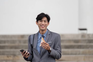 Takım elbise giymiş genç bir işadamı, elinde akıllı telefon ve sandviç tutarken gülümsüyor, öğle yemeği molasını yansıtıyor..