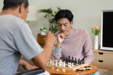 A young man and his father engaging in a chess game at home, showcasing the bond and enjoyment of family activity. clipart