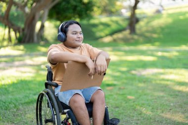 A diverse young man seated in a wheelchair, holding a sketchpad and enjoying music through headphones in a lush green park setting. clipart