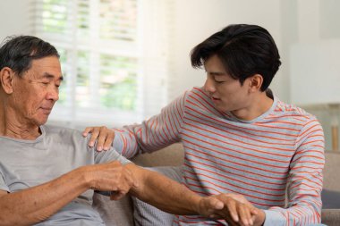 Yaşlı babasını nazikçe koluna dokunarak teselli eden genç bir adam, güçlü bir aile bağı ve ev ortamında duygusal desteği yansıtıyor..