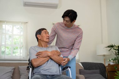 Bir oğul yaşlı babasını tekerlekli sandalyede bağrına basar. Bakıcı bir ev ortamında huzur ve duygusal destek sağlar..