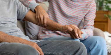 Bir oğul yaşlı babasının elini nazikçe tutar, aile içinde derin bir duygusal bağ ve ilgiyi yansıtır sıcak bir oturma odasında.