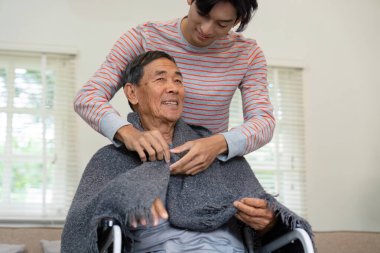 Bir oğul, yaşlı babasına tekerlekli sandalyede otururken battaniyesini düzeltirken yardımcı olur. Rahat bir ev ortamında bakım ve aile desteğini ön plana çıkarır..