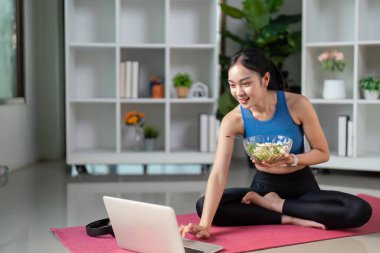 Aktif kıyafetli bir kadın, taze salata yerken dizüstü bilgisayarını kullanırken gülümsüyor ve teknolojiyle besinleri karıştırıyor..