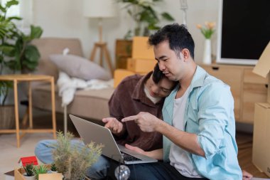 A gay couple sits together, working on a laptop while unpacking boxes in their new home, showcasing teamwork and love. clipart