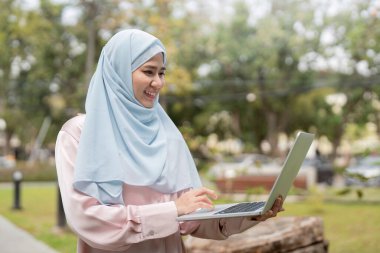 Kendine güvenen, tesettürlü Müslüman bir kadın parkta dizüstü bilgisayarına yazarken gülümsüyor, mesleki bağlılık ve pozitifliği temsil ediyor..