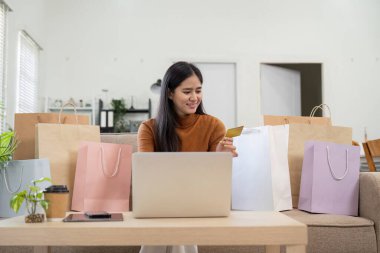 Neşeli bir kadın alışveriş torbalarıyla çevrili kredi kartıyla internetten alışveriş yapıyor..