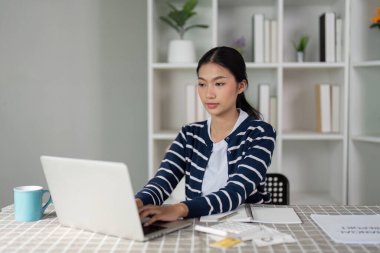 Genç bir kadın finansal planlama ve faturalar için dizüstü bilgisayarda yazıyor. Belgeler ve masanın üzerinde görünür bir defter var..