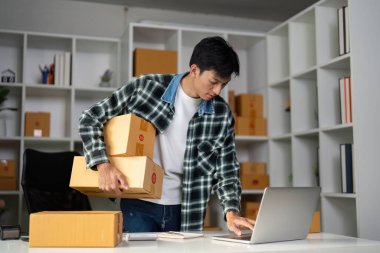 Bir kişi, çevrimiçi satış ve sevkiyatları yönetmek için dizüstü bilgisayar kullanırken bir kolda birkaç karton kutuyu dengeliyor..
