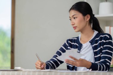 Genç bir kadın mali planlama ve faturalar için notlar yazmaya odaklanmış, bir elinde kalem, diğer elinde çeşitli belgeler tutuyordu..