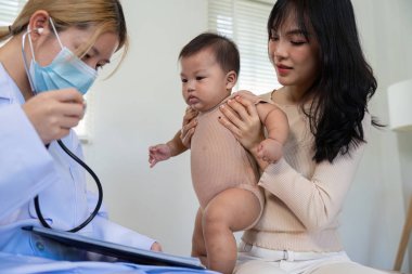 Pediyatri doktoru bebeği muayene etmek için steteskop kullanır ve ebeveyn çocuğu klinik ortamında güvenli bir şekilde tutar..
