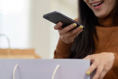 Mutluluk dolu bir kadın alışveriş torbaları tutuyor ve telefonuna gülümsüyor, evdeki online alışveriş deneyimine dalmış..