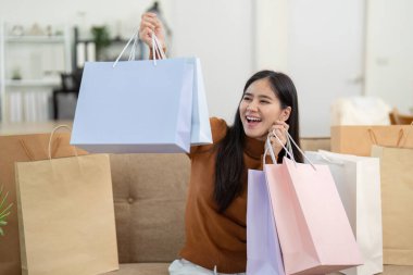 Neşeli genç bir kadın alışveriş çantalarını tutuyor, rahat bir ev ortamında online alışverişlerinin heyecanını dile getiriyor..