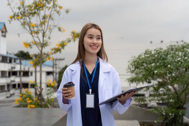 Neşeli genç bir kadın doktor elinde bir kahve ve pano tutarken dışarıda durmuş, dost canlısı bir sağlık hizmetleri ortamını temsil ediyor..