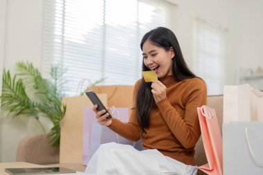 Keyifli bir kadın telefonunu ve alışveriş çantalarını tutarken gülümsüyor, online alışverişlerini sıcak bir ortamda kutluyor..