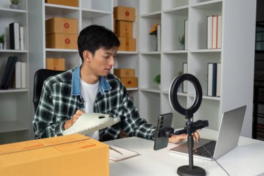 Genç bir girişimci, bir ürünü sergilerken çevrimiçi satışları yönetirken, bir dizüstü bilgisayar ve akıllı telefonu kutularla dolu modern bir çalışma alanında kullanıyor.