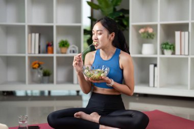 Bir kadın, yemeğinden gülümseyerek, yoga minderinde rahatça oturarak, sağlıklı bir yaşam tarzına ve kendine özen göstererek zevk alır..
