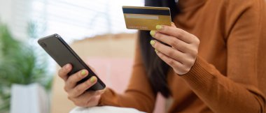 Genç bir kadın, lüks yaşam alanında alışveriş torbalarıyla çevrimiçi alışveriş yaparken dizüstü bilgisayarını ve kredi kartını kullanıyor..